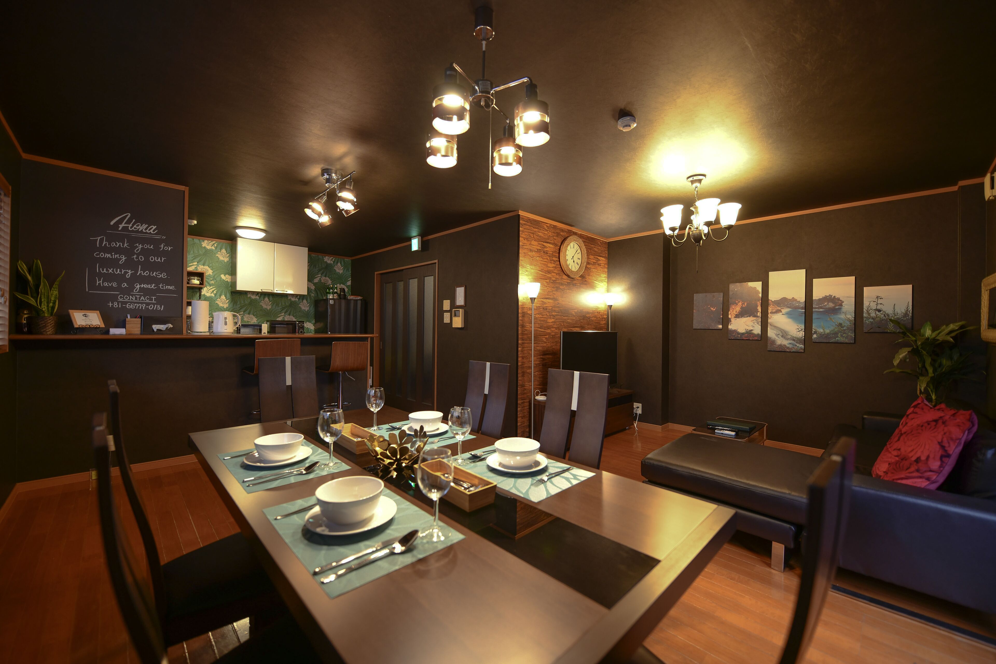Un comedor con una larga mesa dispuesta para cuatro personas, un área de estar separada con un sofá, y una cocina visible al fondo.