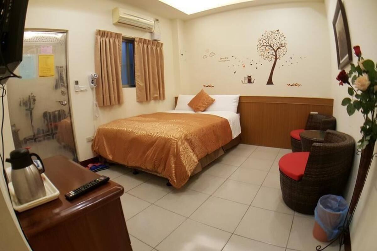 A hotel room with a bed, a desk with a coffee maker, a chair, a small table, and a wall-mounted TV.