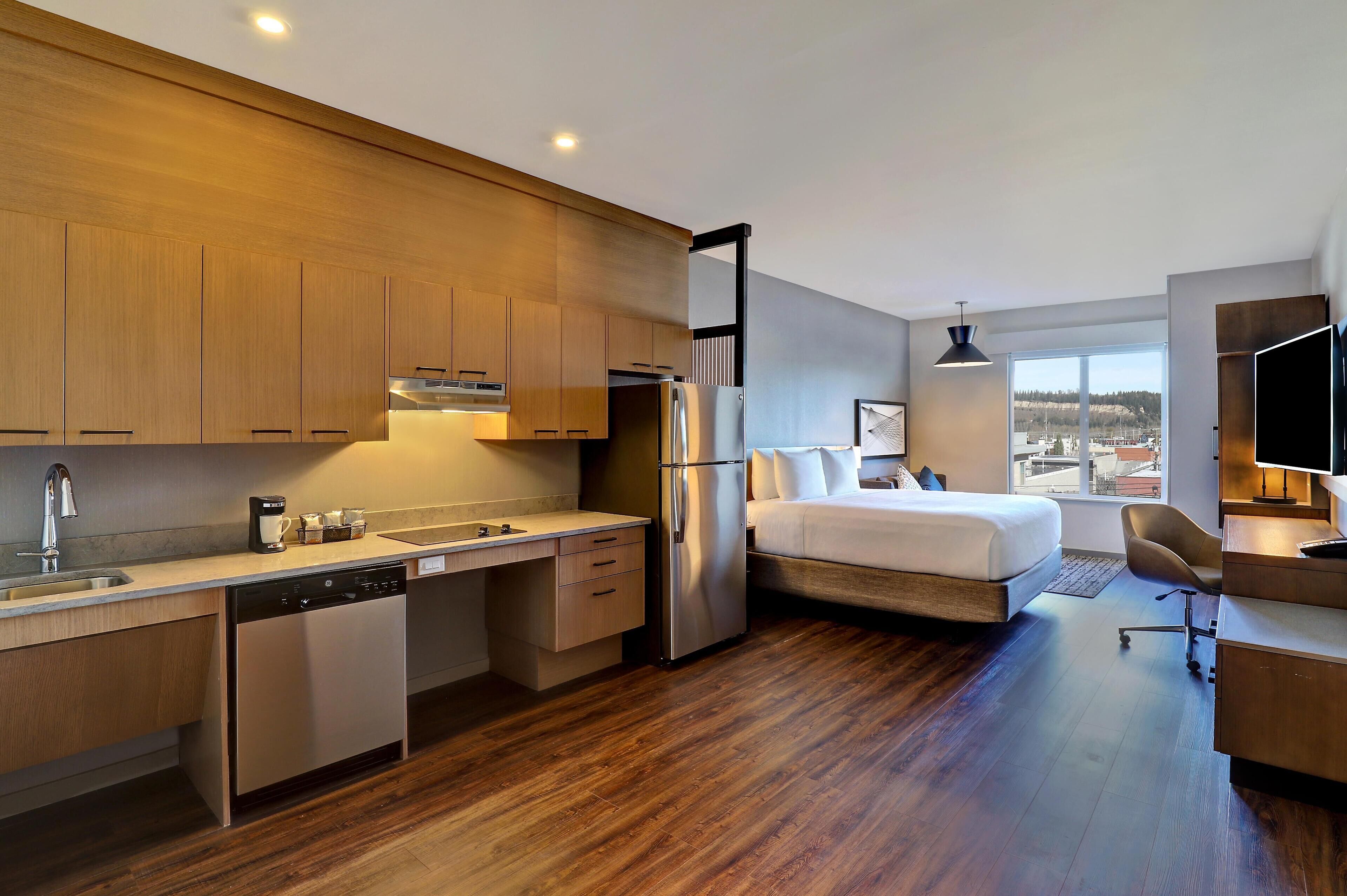 Ein modernes Hotelzimmer mit Küchenzeile, einem Bett, einem Schreibtisch und Blick auf die Stadt.