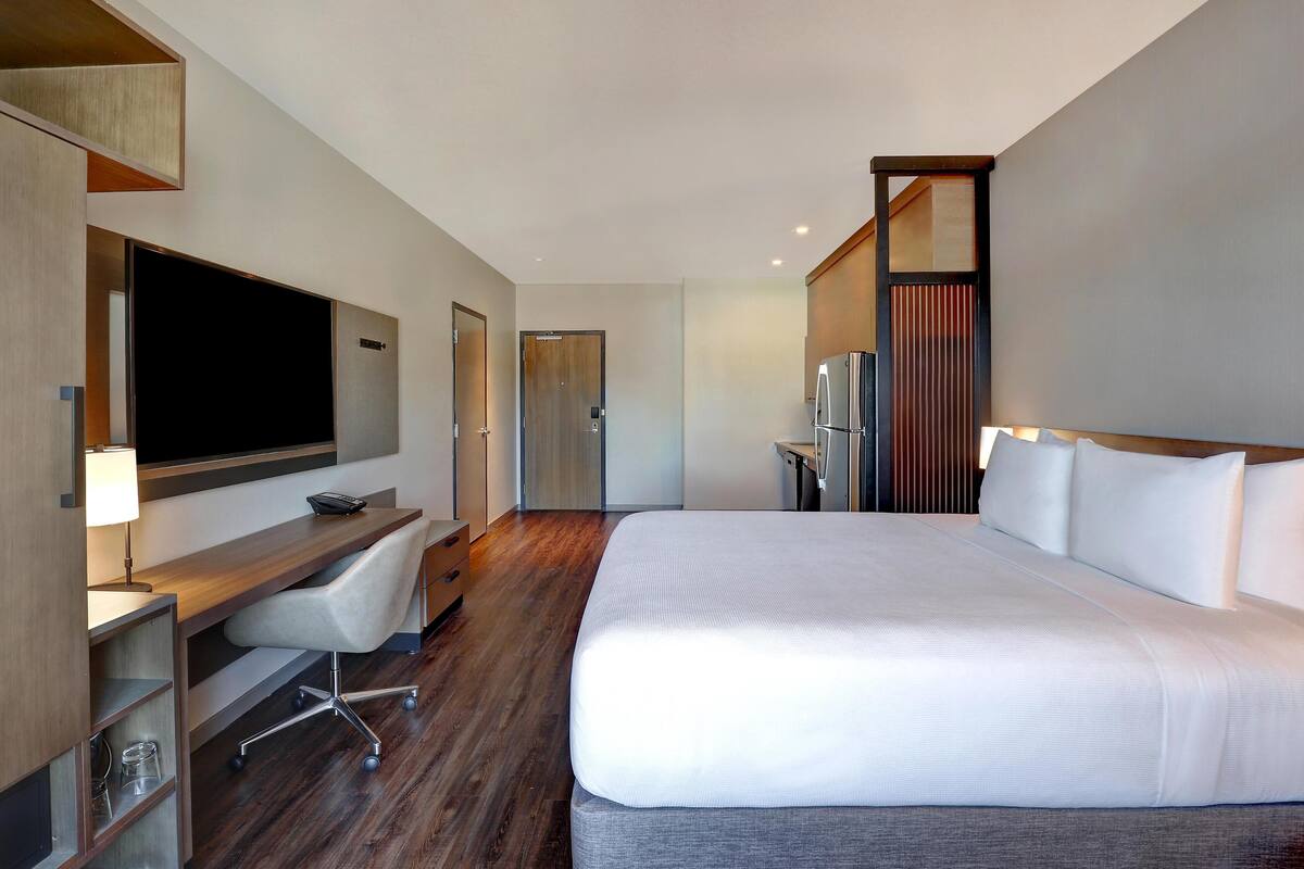A modern hotel room with a large bed, a desk with a chair, a flat-screen TV, and a refrigerator.