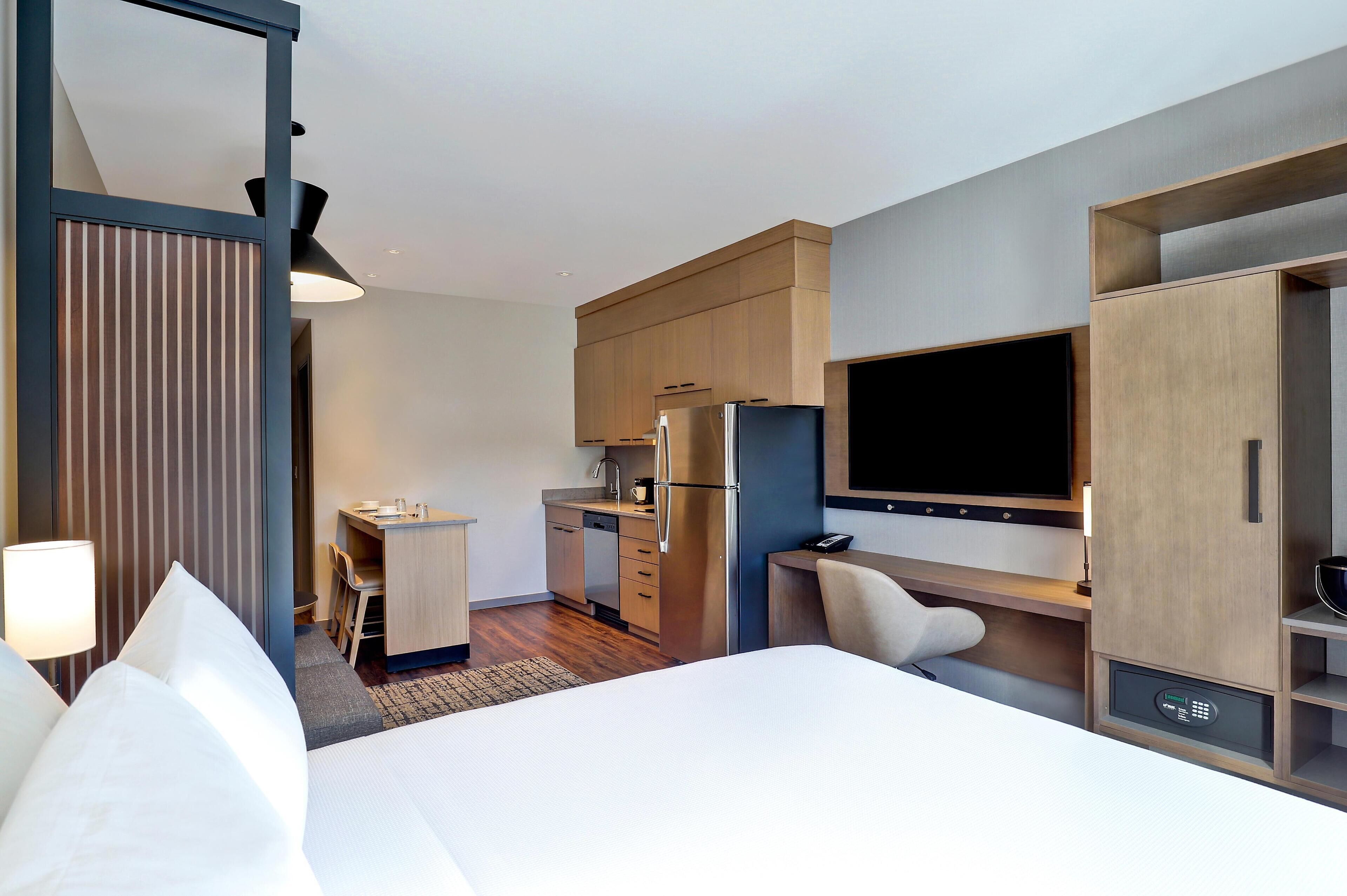A modern hotel room with a bed, a desk, a kitchenette, and a TV.