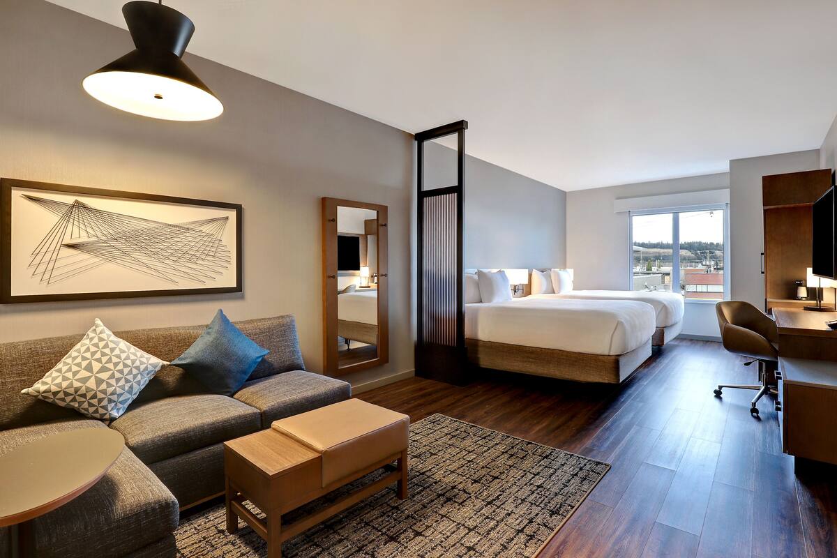 A modern hotel room with a large bed, a sofa, a desk, and a large window offering a city view.