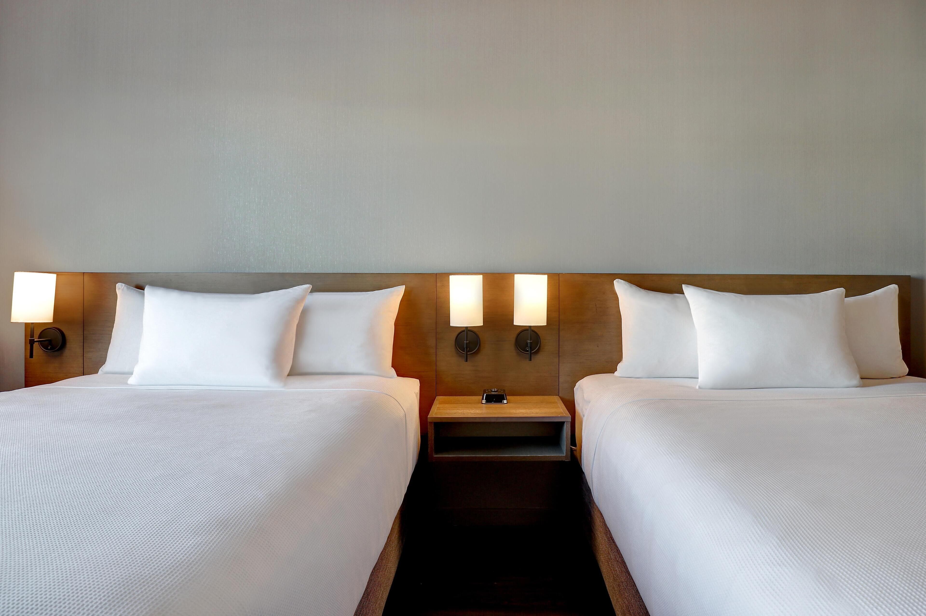 Habitación de hotel con dos camas, cabecero de madera, lámparas de noche y mesitas auxiliares.
