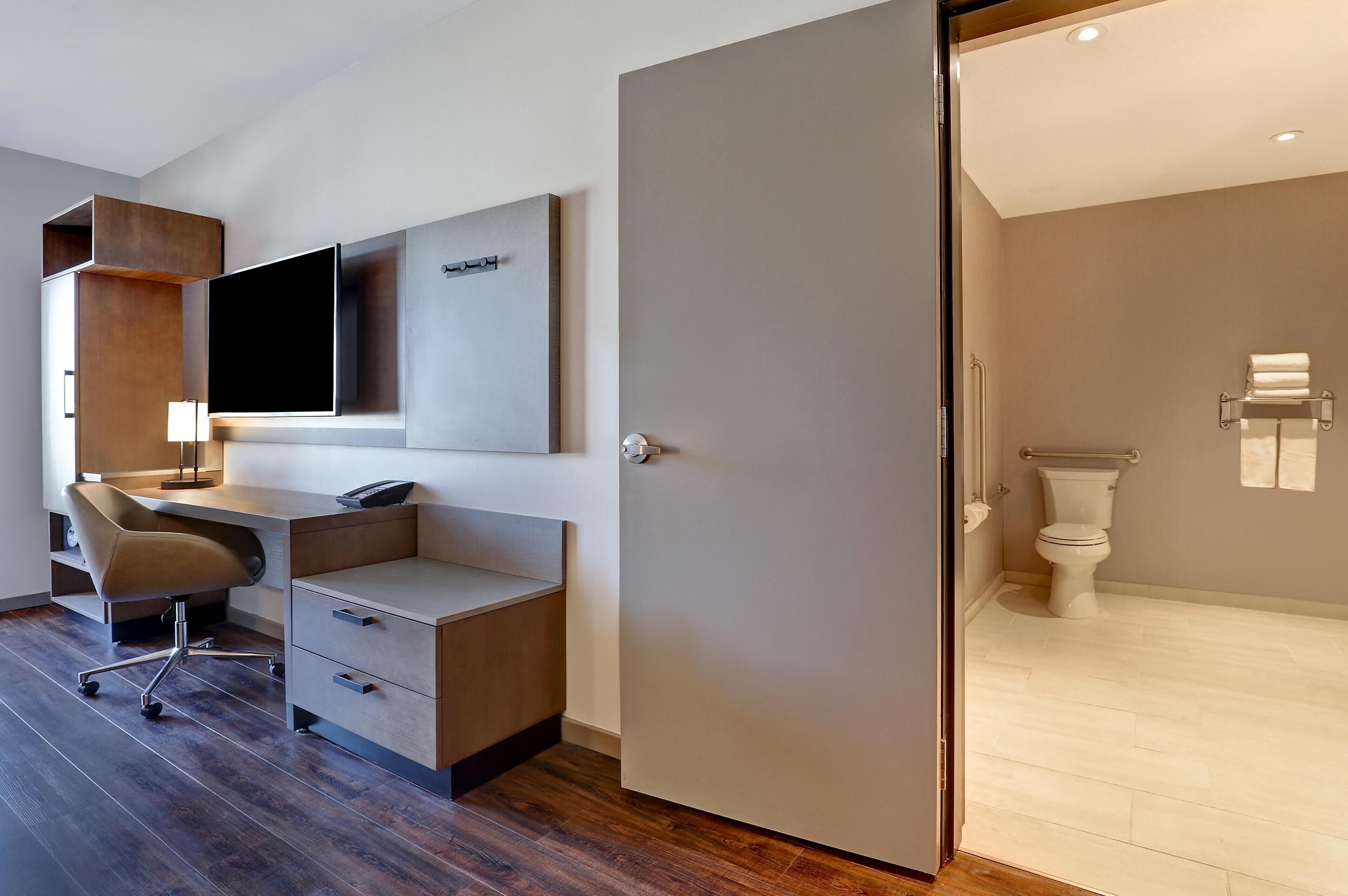 Ein modernes Schlafzimmer mit einem großen Bett, einem Schreibtisch mit Stuhl, einem Flachbildfernseher und einem Badezimmer mit Toilette und Handtuchhalter.