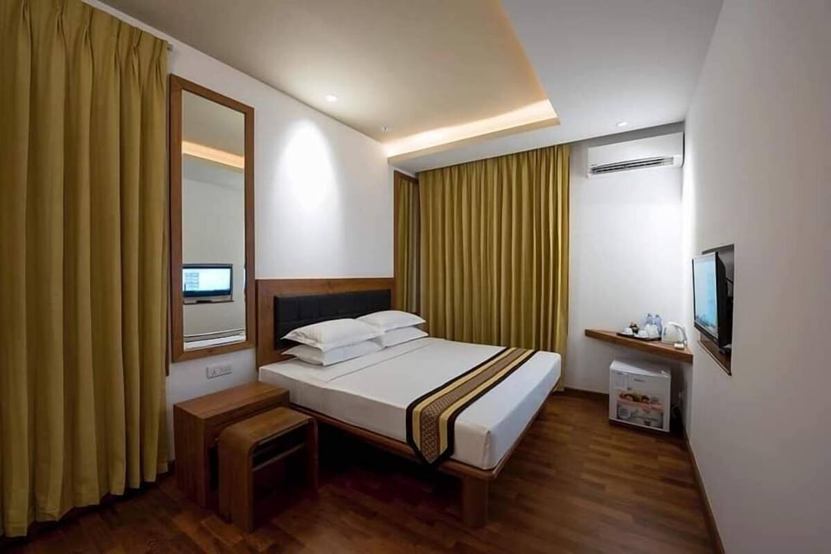 A modern hotel room with a bed, bedside table, mirror, curtains, TV, and a small cabinet.