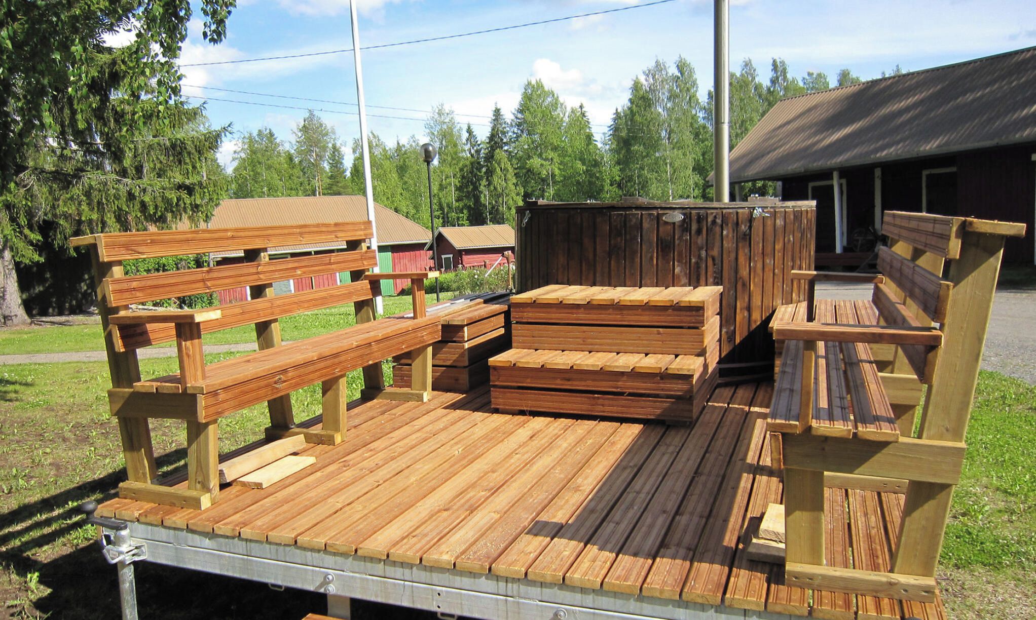 A wooden deck with benches and a table, surrounded by trees and a building in the background.