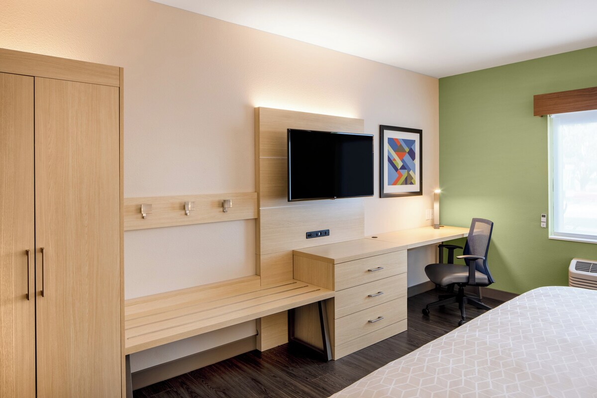 Quarto moderno com cama, escrivaninha e cadeira. Há uma televisão na parede e um quadro acima da escrivaninha.