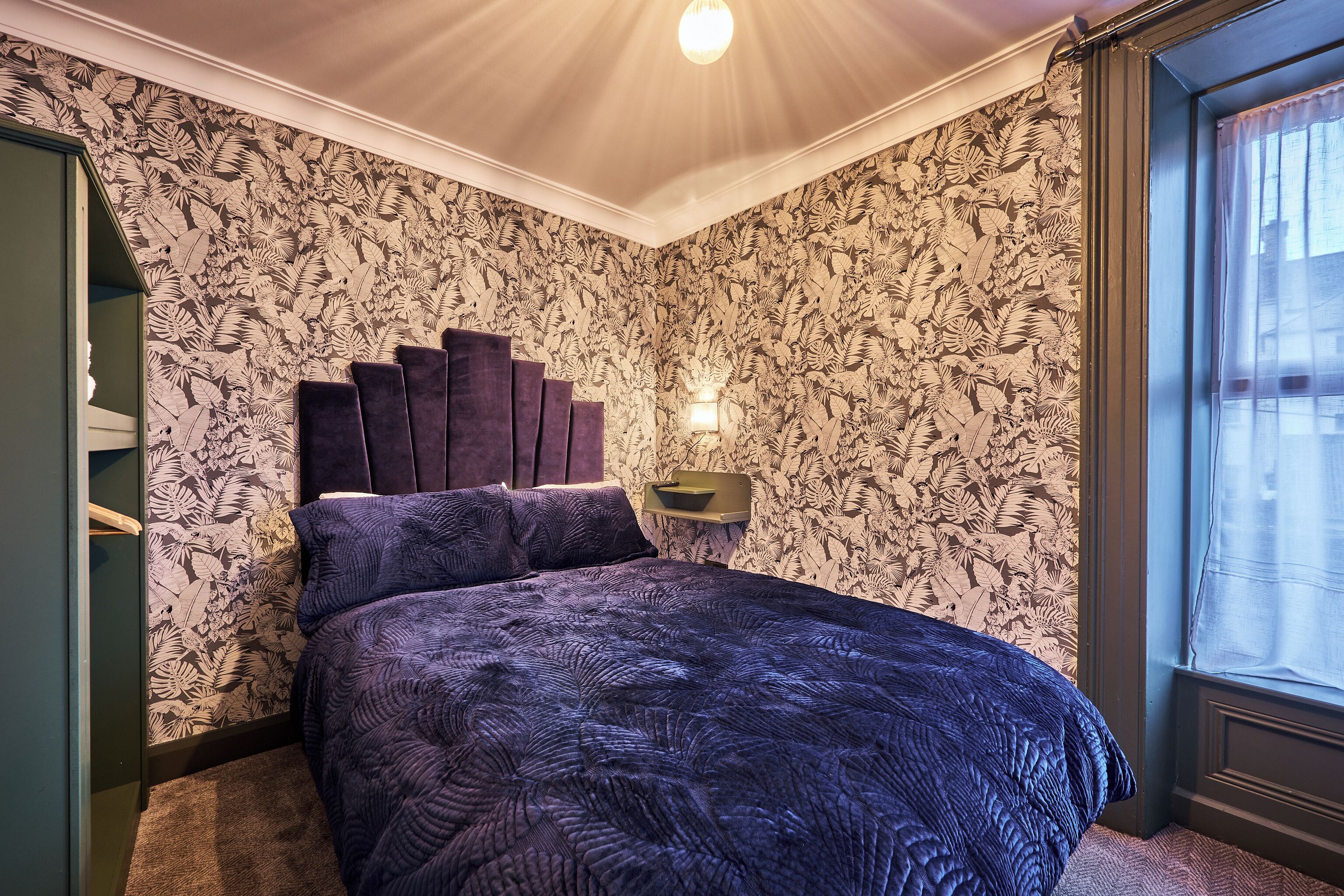 Un dormitorio con cama, mesita de noche, luz empotrada en la pared y ventana con cortinas.