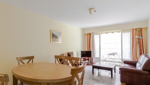 Een woonkamer met een ronde houten tafel, diverse stoelen, een bank, een televisie en een balkon met gordijnen.