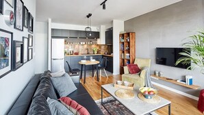 A modern living room with a sofa, coffee table, and a TV. A kitchen is visible in the background with a dining area. - Elegant Apartment in Zoliborz | Balcony and Parking (Warszawa)