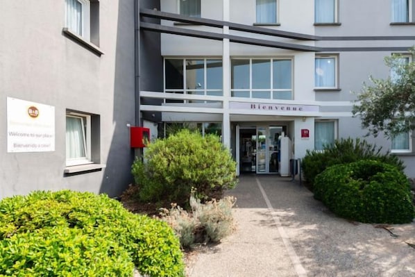 A building with a sign reading 'Bienvenue' at the entrance, a red fire alarm box, and a gravel pathway leading to the door. - B&B Hotel Montauban (Montauban)