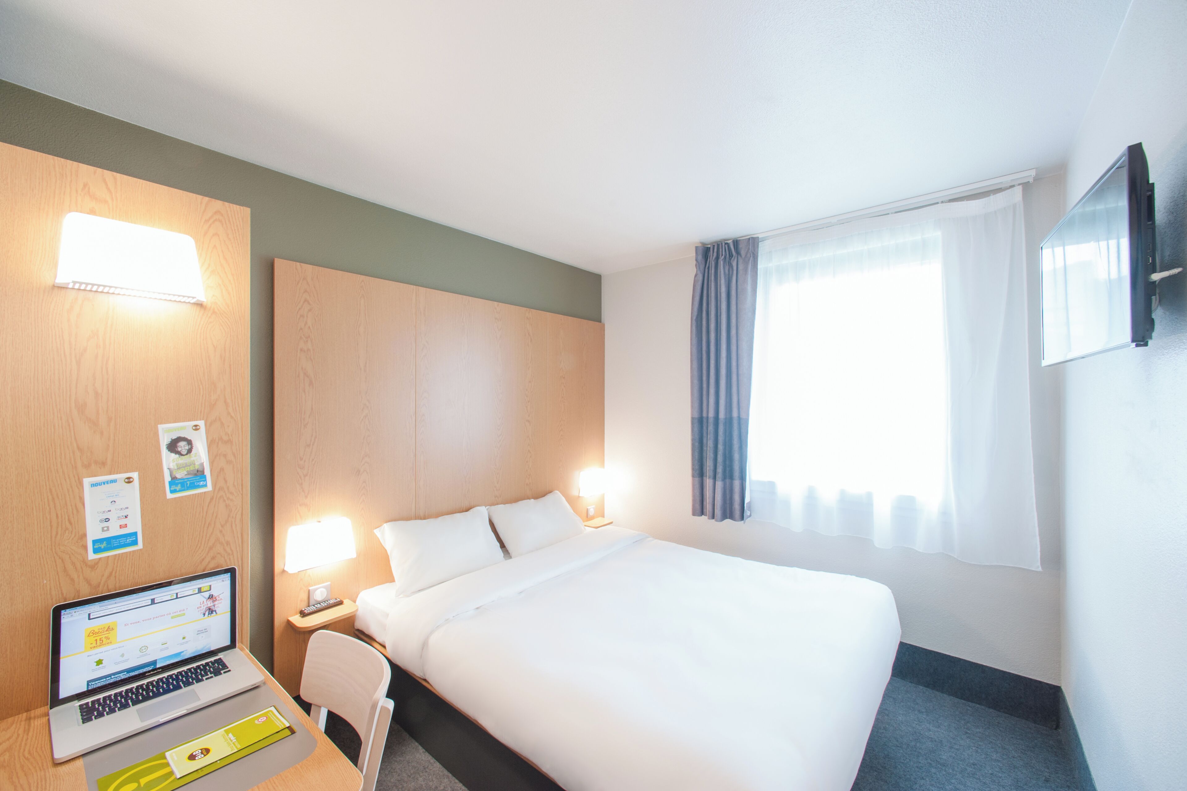 Une chambre d’hôtel avec un lit, un bureau avec un ordinateur portable, une lampe et un téléviseur fixé au mur.