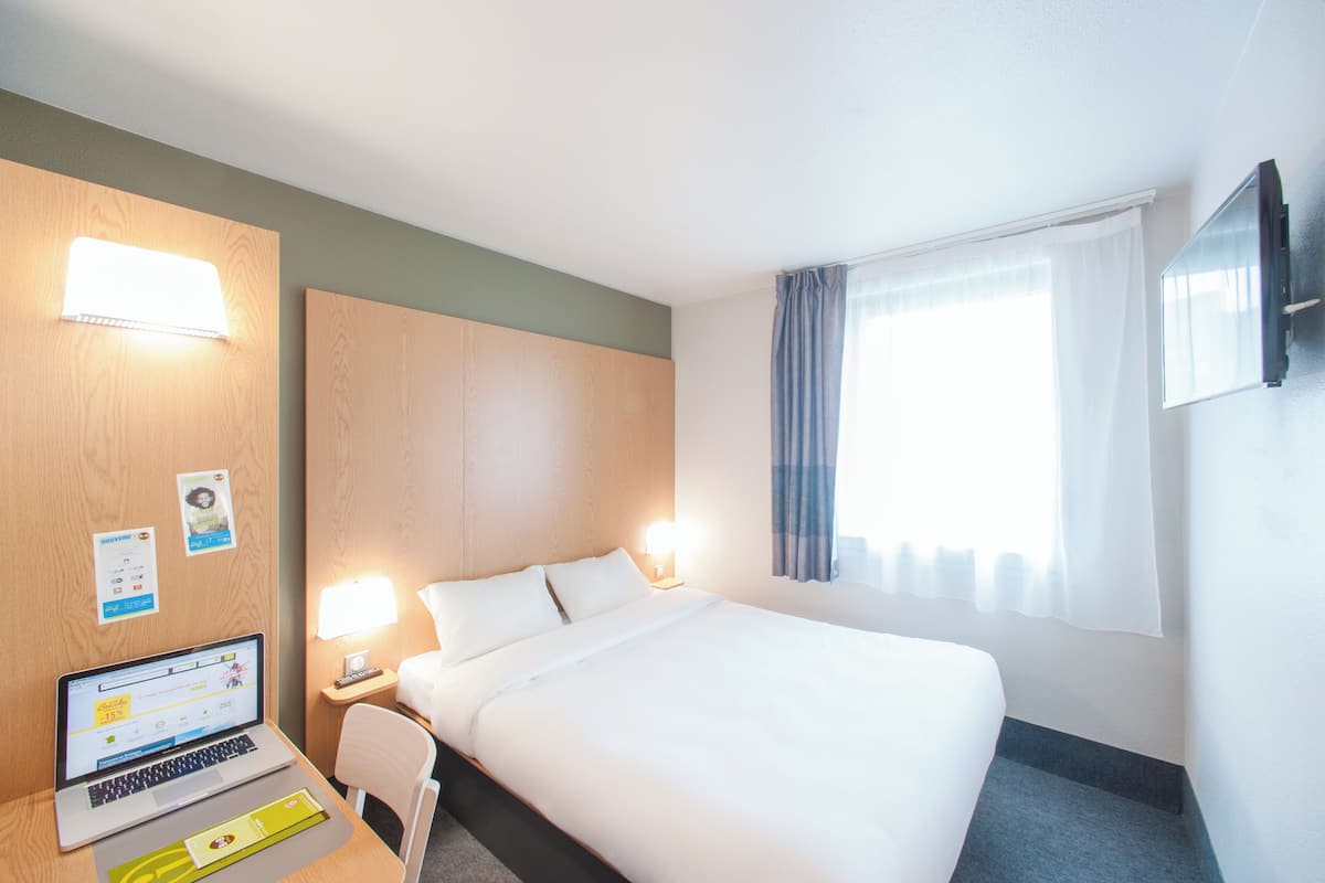 Une chambre d’hôtel avec un lit, un bureau avec un ordinateur portable, une lampe et un téléviseur fixé au mur.