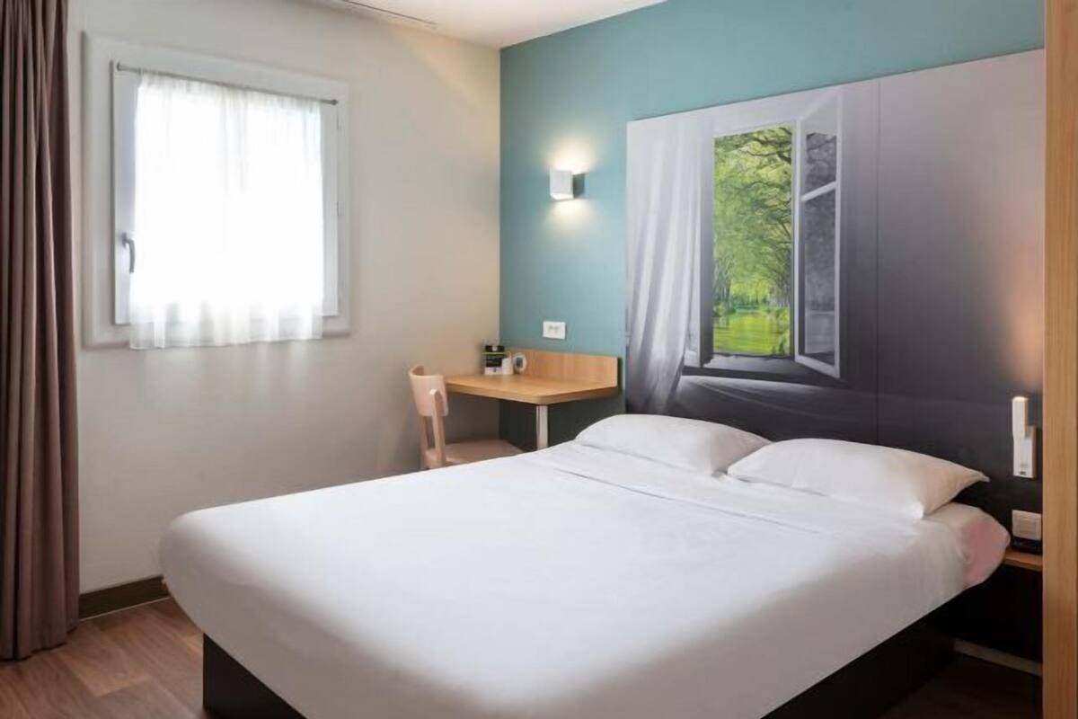 Une chambre d’hôtel équipée d’un lit, d’un bureau, d’une chaise, d’une fenêtre avec des rideaux et d’une lampe fixée au mur.