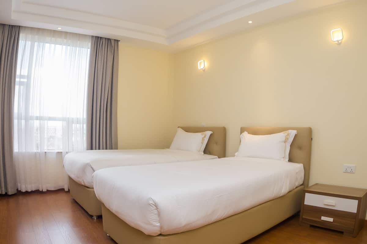 A hotel room with two beds, a wooden floor, a window with curtains, and a nightstand with a drawer.