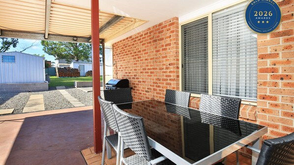 Eine Terrasse im Freien mit einem Glastisch und Stühlen, einem Grill und einer Ziegelwand.