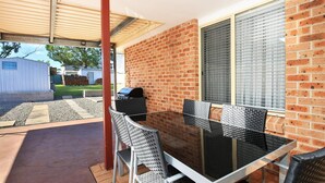 Un patio con mesa y sillas de vidrio, un muro de ladrillo y una parrilla.