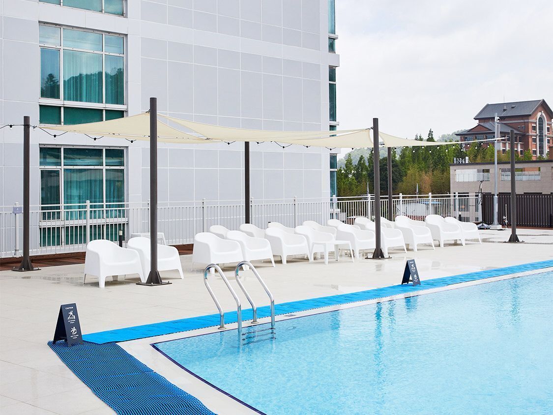Une piscine entourée de chaises longues blanches et d’un espace ombragé, le tout bordé d’un bâtiment blanc doté de grandes fenêtres.
