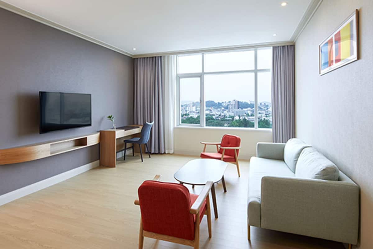 Une chambre d’hôtel moderne dotée d’un téléviseur à écran plat fixé au mur, d’un bureau avec une chaise, d’un canapé et d’une grande fenêtre donnant sur la ville.