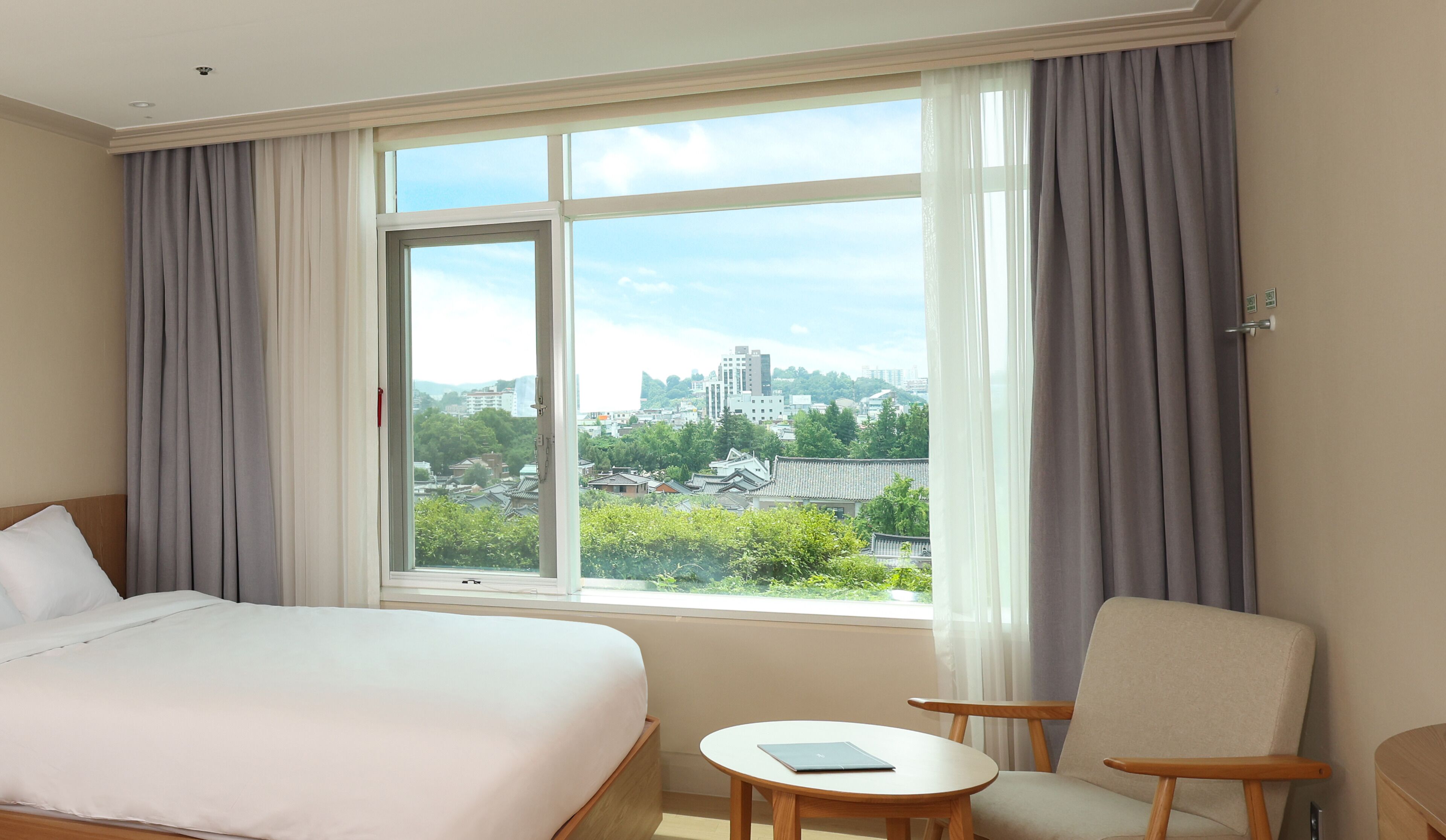 A hotel room with a large bed, a small table, a chair, and a window with a city view.