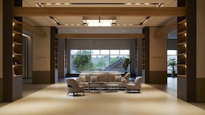 Een ruime lobby met een modern ontwerp, met een centraal zithoek met een bank en fauteuils, een groot raam met uitzicht naar buiten, en een boekenkast aan de linkerkant.