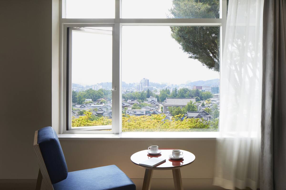 Une pièce avec une grande fenêtre, une chaise, une petite table avec deux tasses et un livre.