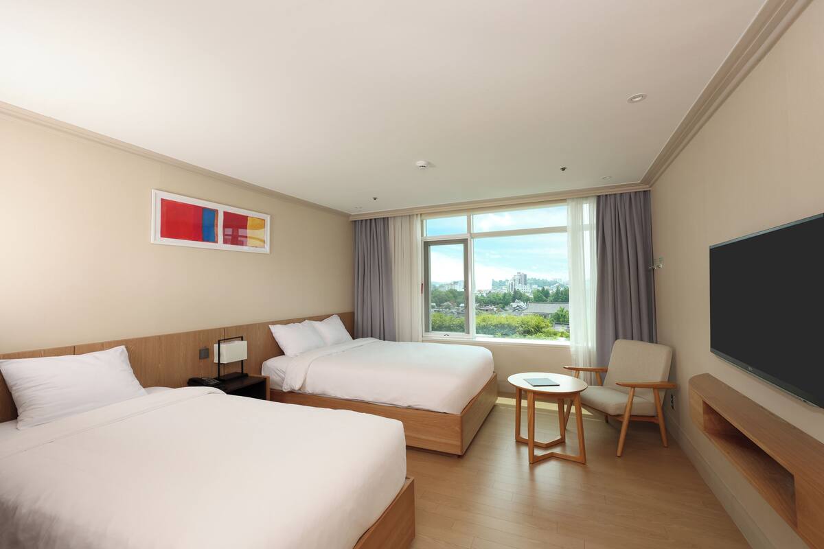 Une chambre d’hôtel avec deux lits, un bureau, une chaise, une télévision et une grande fenêtre donnant sur la ville.