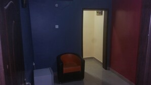 A narrow hallway with a blue wall, a red wall, and a white door at the end. There is a small white appliance on the left and a black and orange chair in the center.