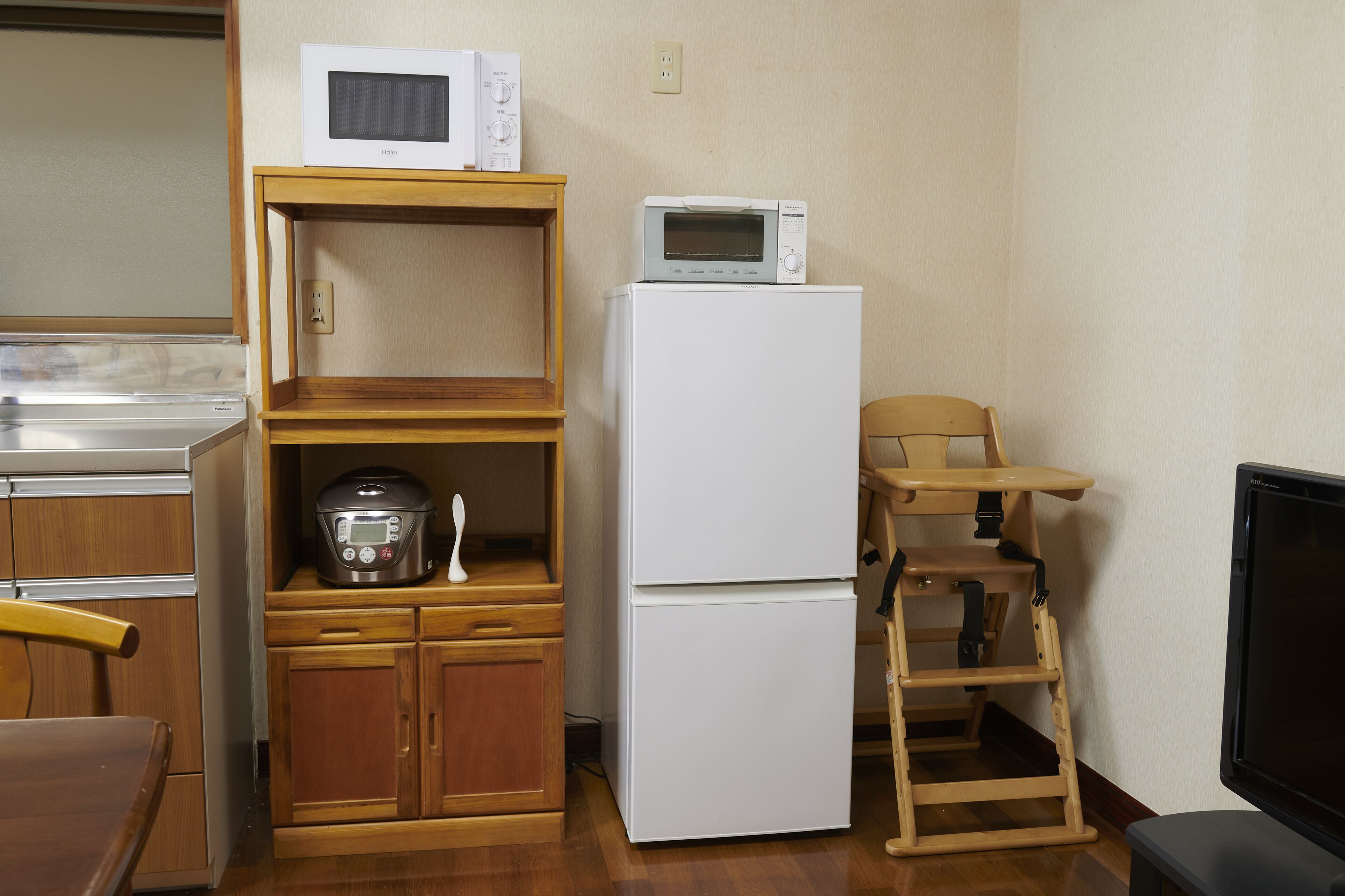 A compact kitchen with a microwave, a refrigerator, a wooden cabinet, a chair, and a television.