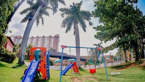 Un parque infantil con toboganes y columpios, rodeado de palmeras y un edificio al fondo.