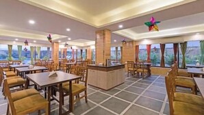 A spacious dining area with multiple tables and chairs, a central counter, and large windows offering a view of the outdoors. - LBD Resorts & Hotels (Kolkata)