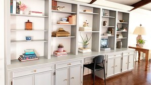 Una habitación amplia con un mueble blanco y alargado con varios estantes, una mesa de madera con una lámpara y una silla negra.