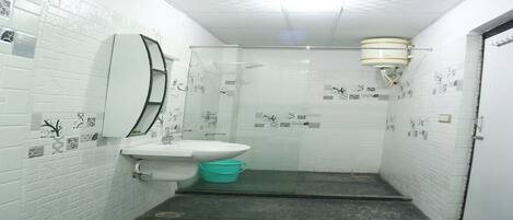 Een kleine, smalle badkamer met een wit wastafel, spiegel en douche. De muren zijn betegeld met een wit patroon, en er staat een groene prullenbak.