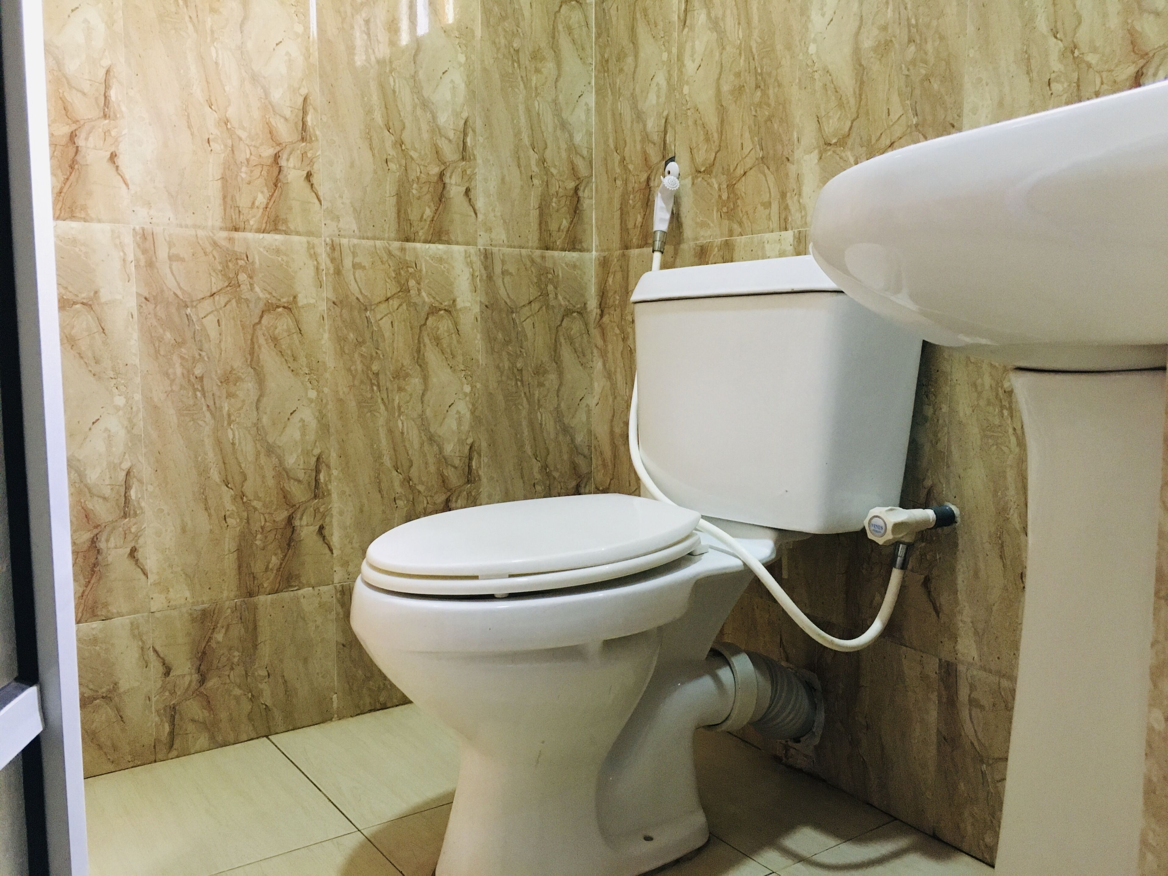 Des toilettes avec une cuvette et un réservoir blancs, reliées à un tuyau fixé au mur, dans une salle de bain aux murs en marbre et au sol carrelé.