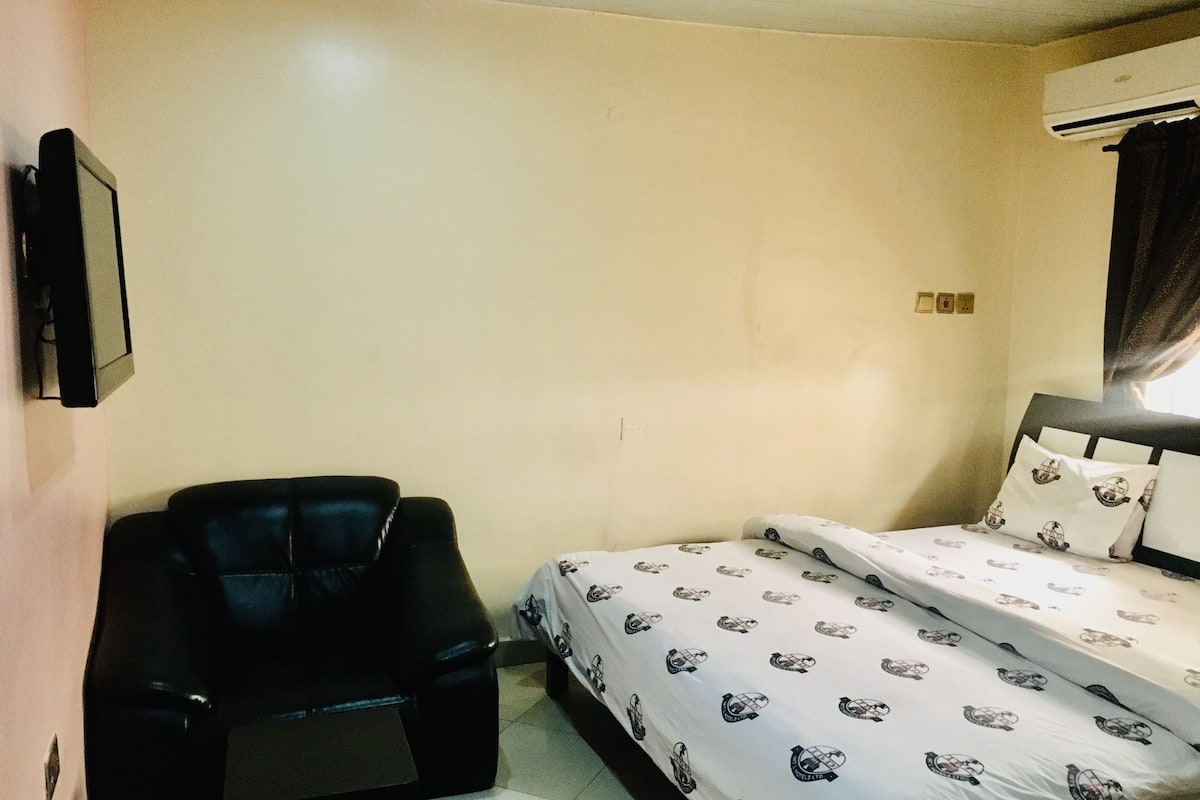 A small, simple room with a bed, a chair, and an air conditioning unit.