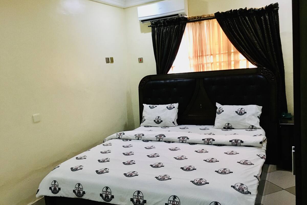 A single bed with a headboard, a window with sheer curtains, and an air conditioning unit on the wall.