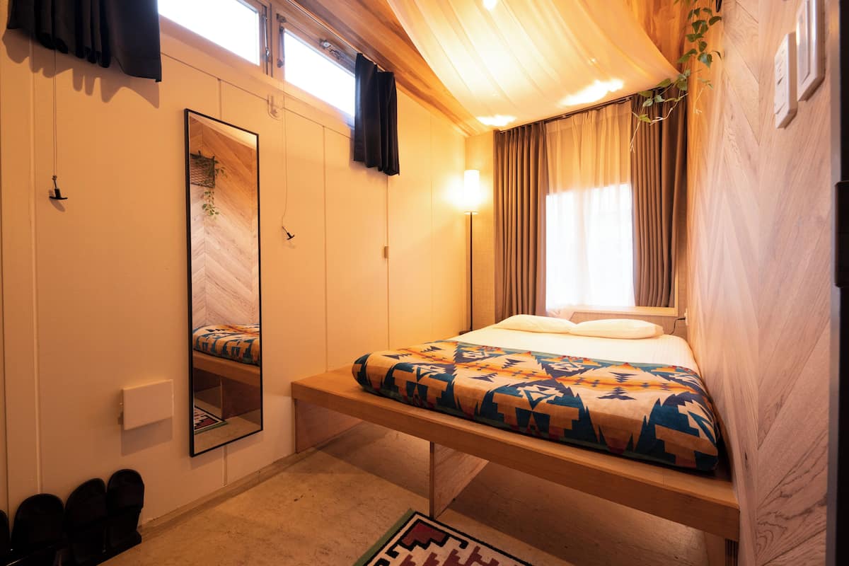 Un dormitorio con cama, espejo, mesita de noche, lámpara y ventana con cortinas.
