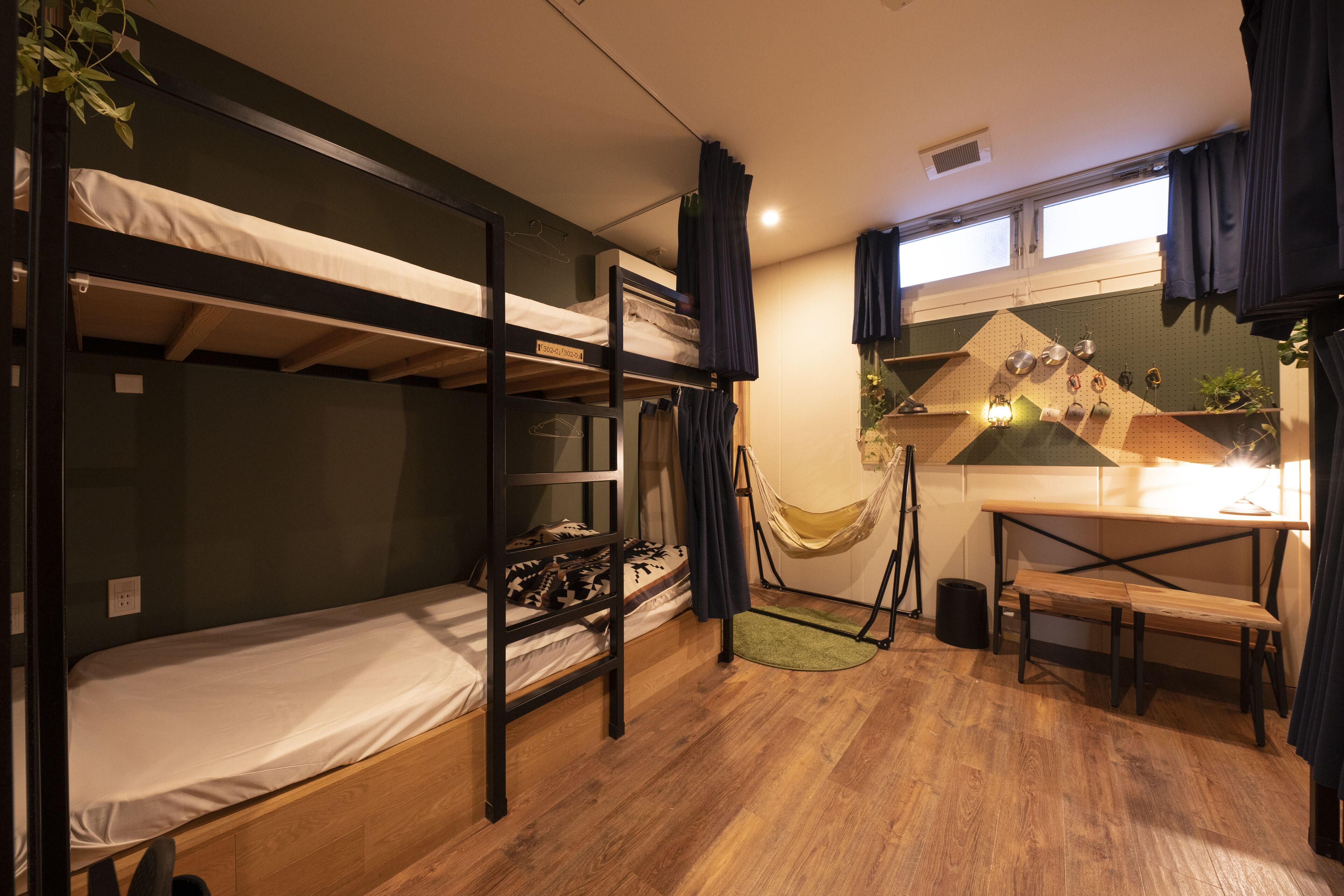 A dormitory room with bunk beds, a desk, a hammock, and a pegboard wall with kitchen utensils.