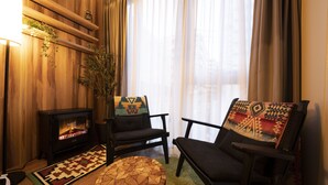 Un salon chaleureux avec un poêle à bois, deux fauteuils, une table ronde en bois et une grande fenêtre ornée de rideaux légers.