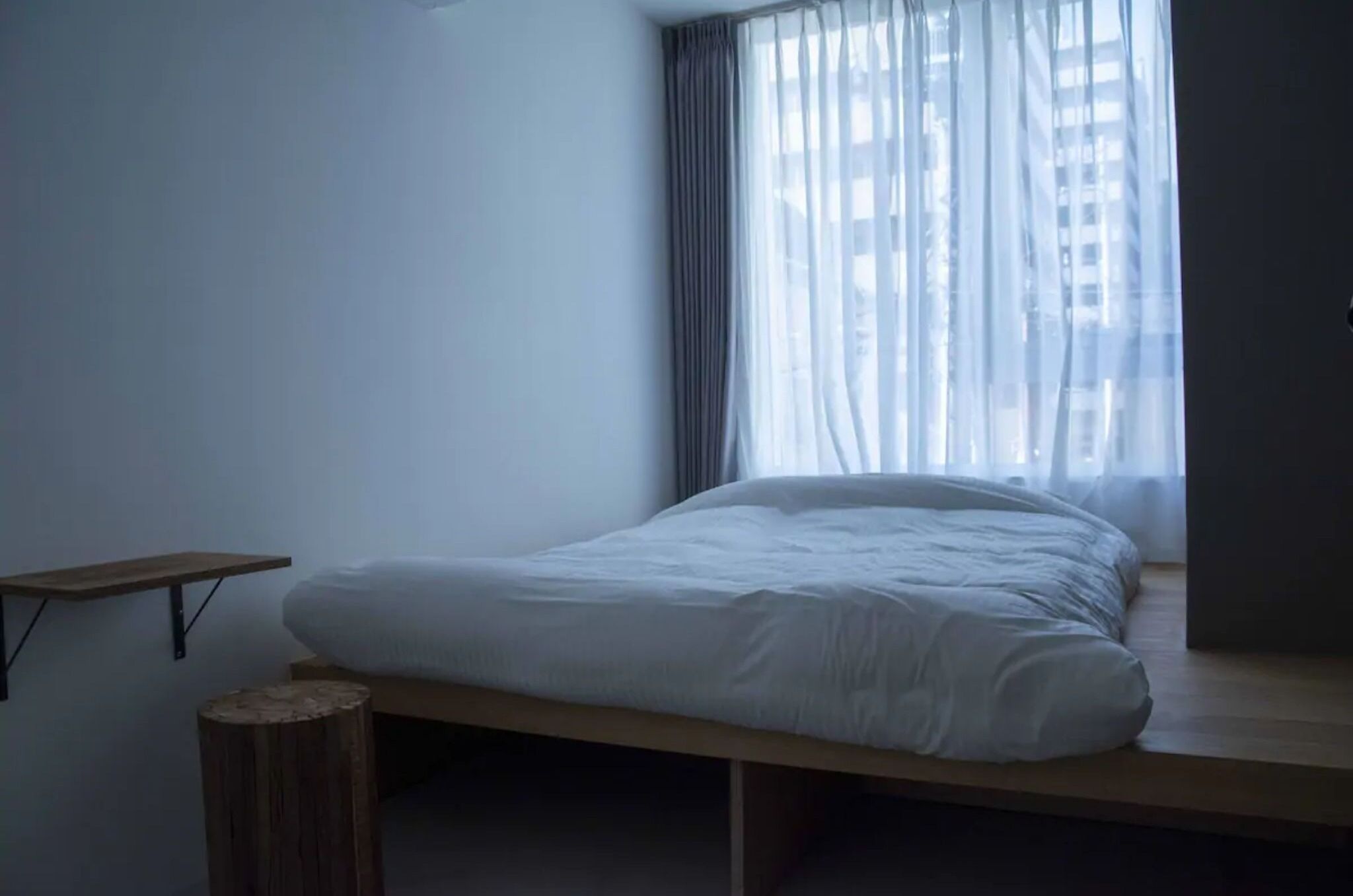 Ein schlicht eingerichtetes Schlafzimmer mit einem Einzelbett, einem kleinen Holzschemel und einem Regal.