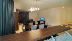 A modern living room with a wooden dining table, black and white chairs, a flat-screen TV, and a ceiling light fixture. - The Capital (Abuja)