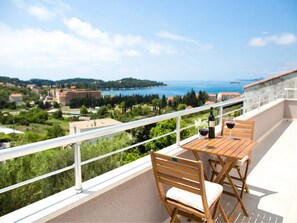 Un balcon avec une table et des chaises en bois, une bouteille de vin et un verre de vin. Le balcon offre une vue panoramique sur une zone côtière, avec des bâtiments et de la végétation.