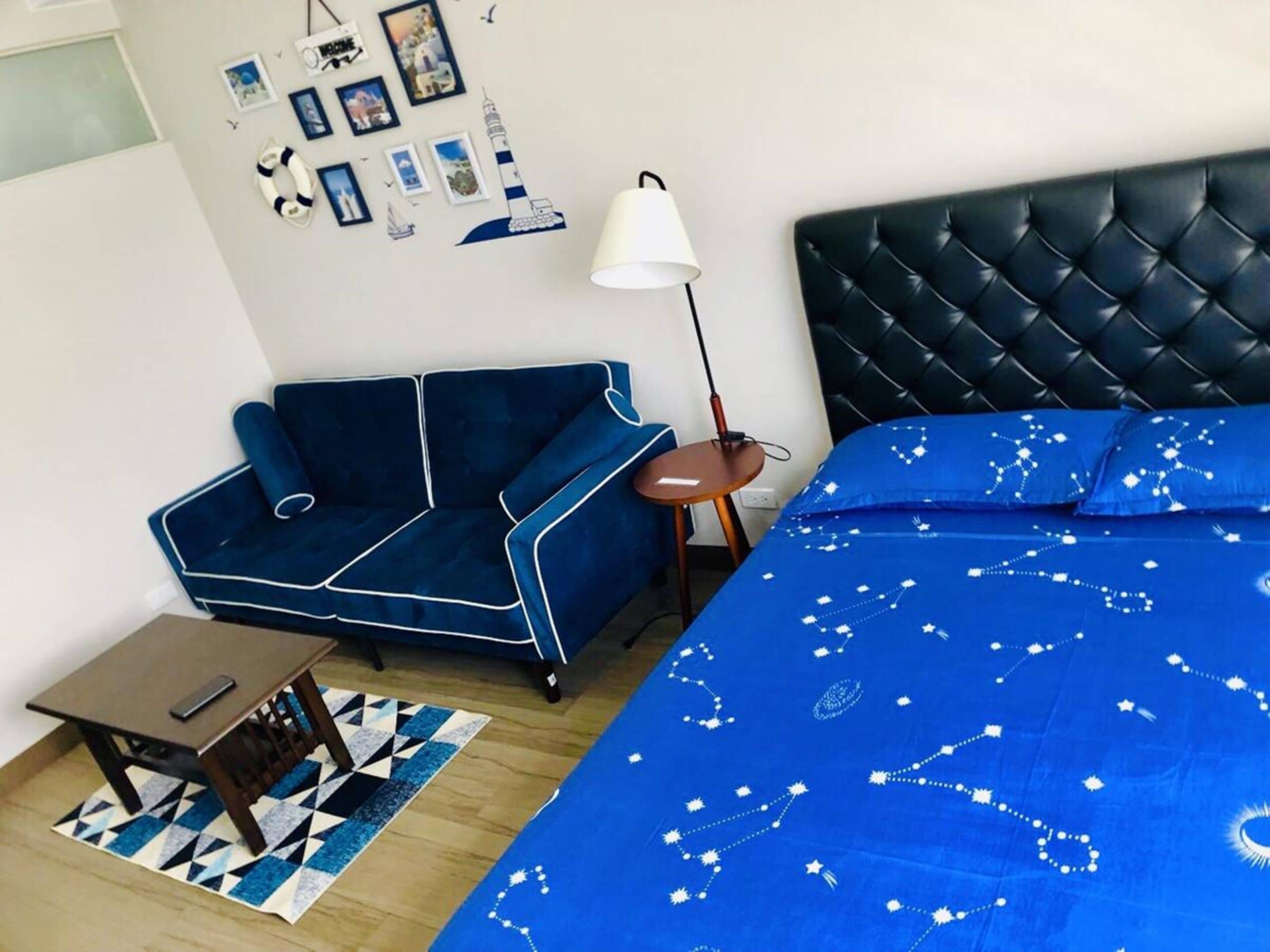Una habitación con un sofá azul, una cama con colcha azul, una mesita con lámpara y una pared decorada con cuadros y un flotador.