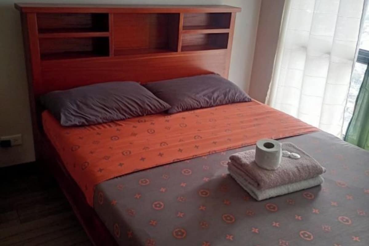 A bed with a patterned bedspread and pillows, a wooden headboard, and a window with curtains.