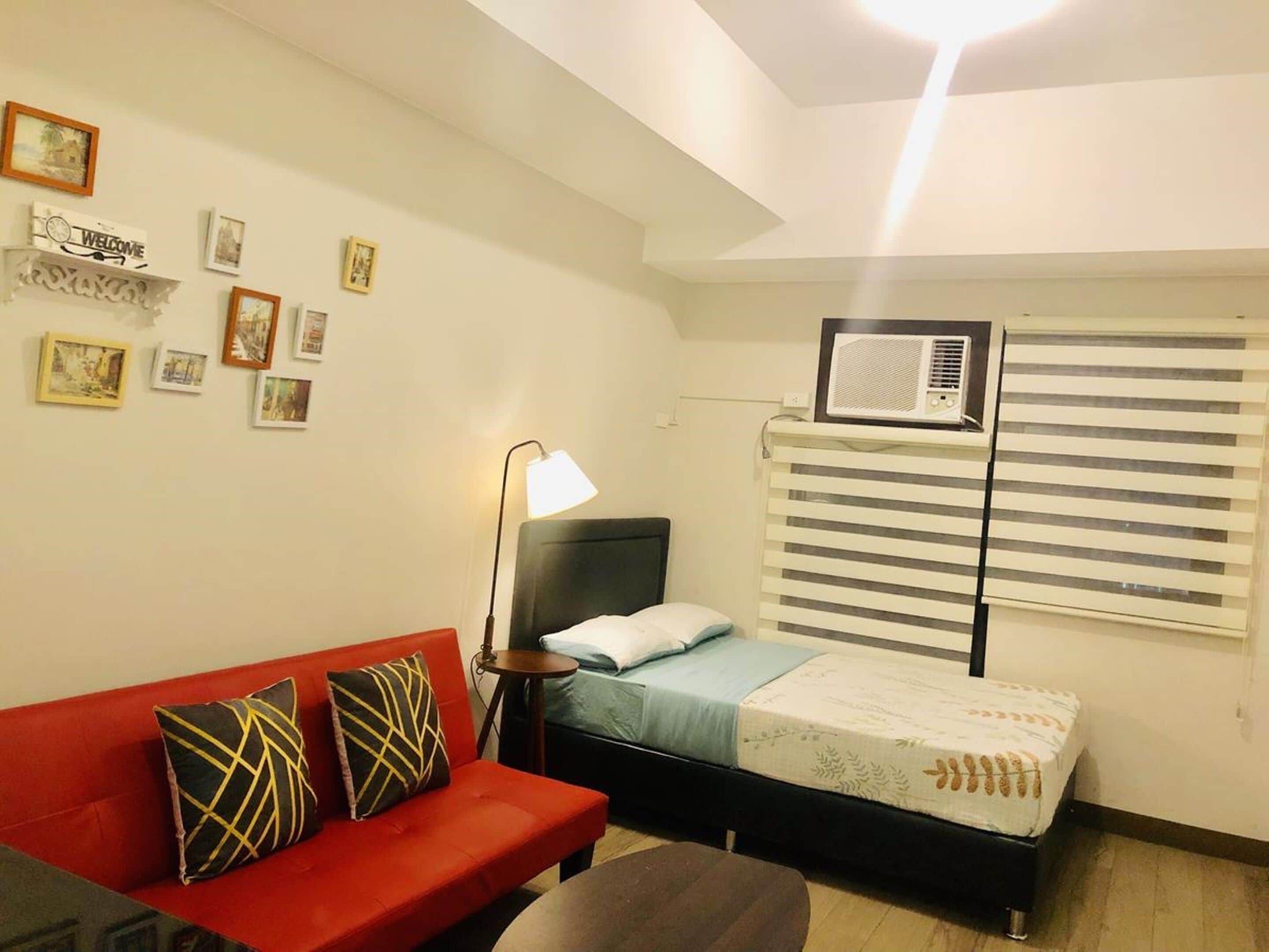 A small, well-lit room with a single bed, a red sofa with patterned cushions, a bedside table with a lamp, and a wall-mounted air conditioner.