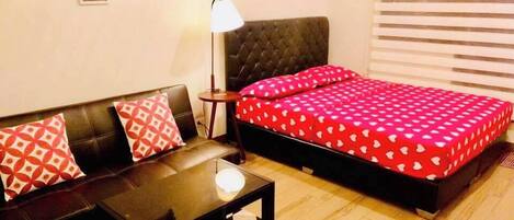 A room with a bed, a sofa, a small table, and a lamp.