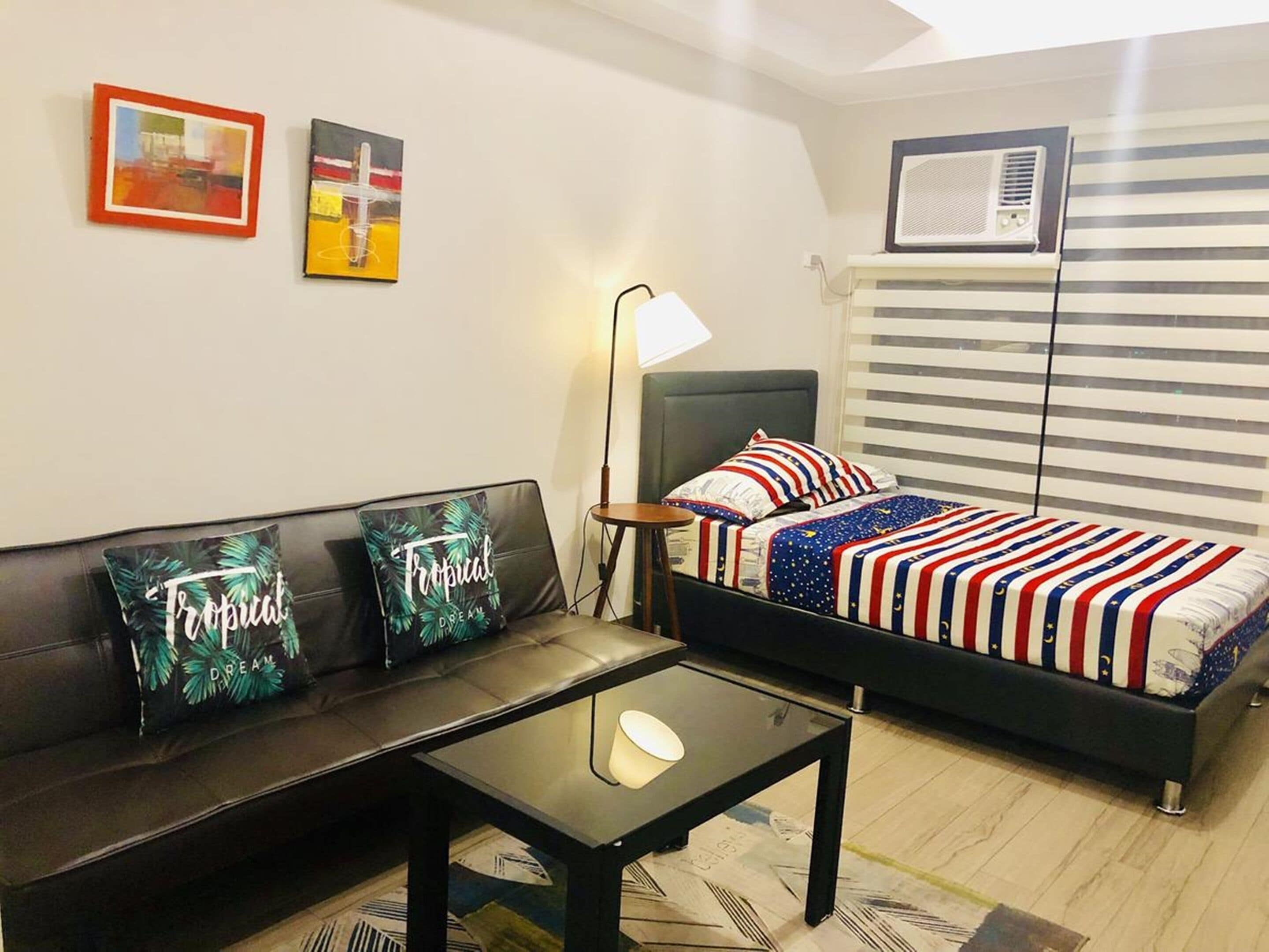A room with a bed, a sofa, a small table, and a nightstand. There are two wall-mounted air conditioning units and a window with blinds.