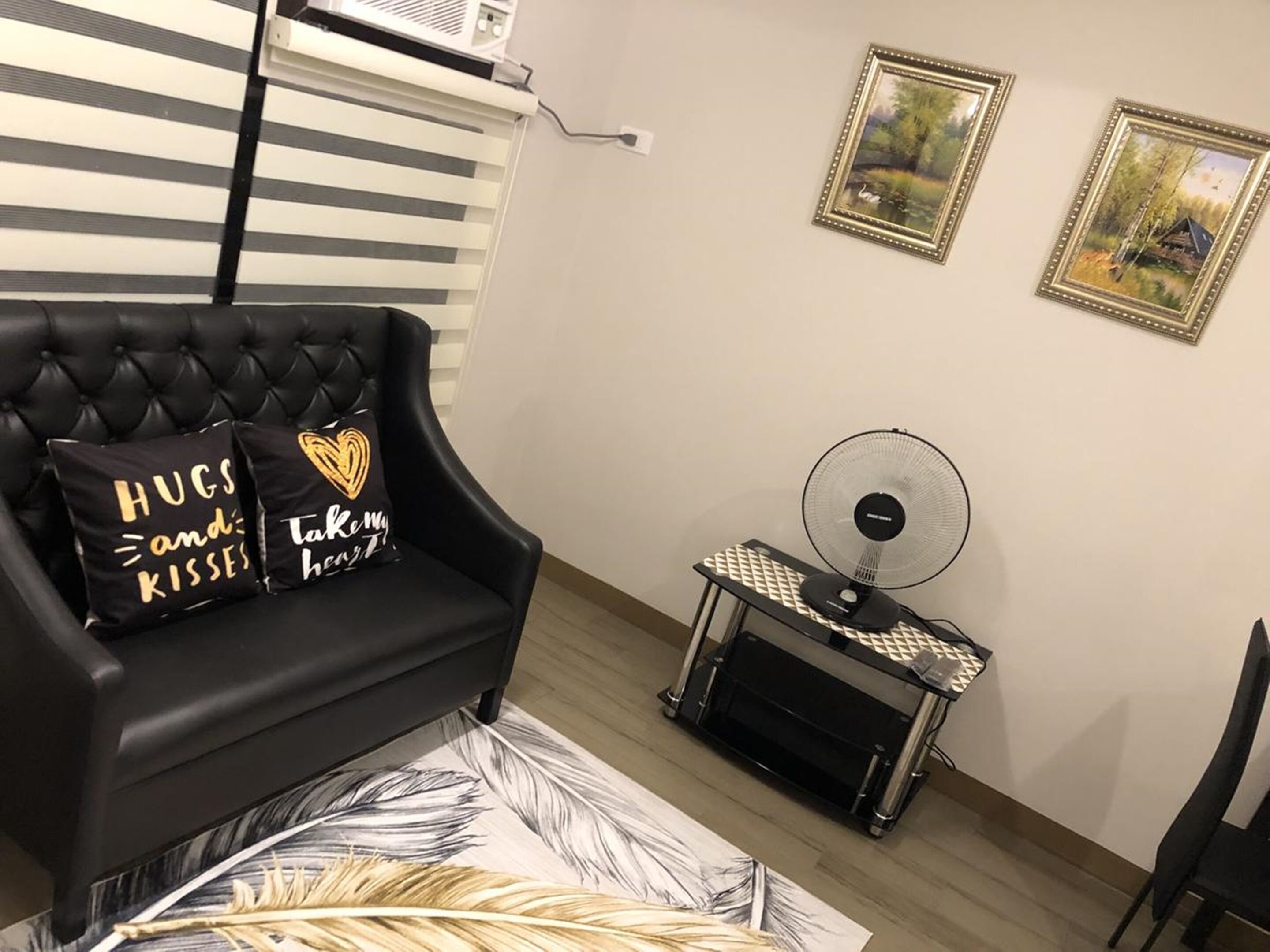 A room with a black leather sofa, two pillows with text, a standing fan, and two framed pictures on the wall.