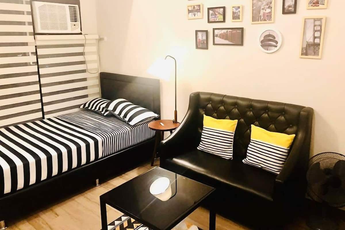 A room with a striped bed, a black leather sofa with yellow pillows, a small table, and a wall with framed pictures.