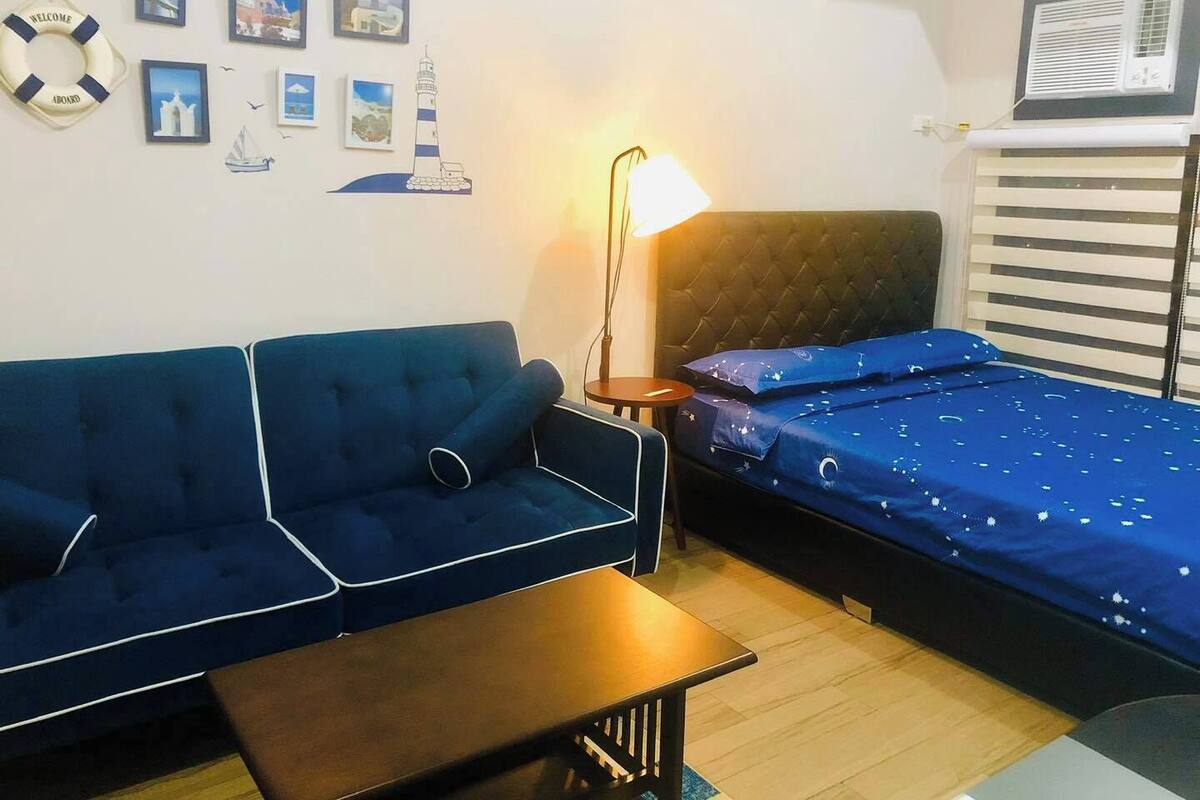 A room with a blue sofa, a small table, a bed with a blue bedspread, a wall-mounted air conditioner, and various framed pictures on the wall.